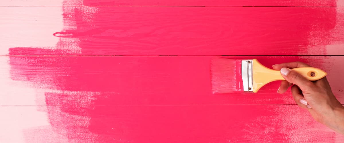 Hand using a paintbrush to apply bright pink paint onto a wooden surface