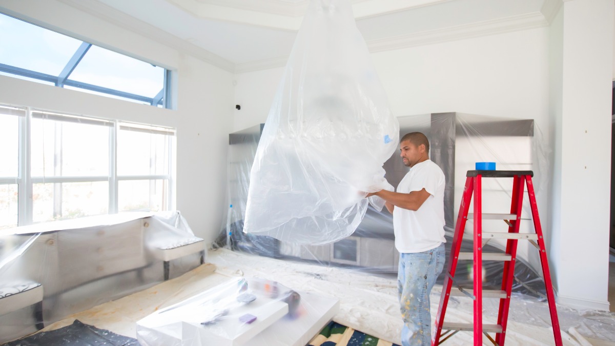 A painter covering a hanging chandelier with a plastic drop cloth
