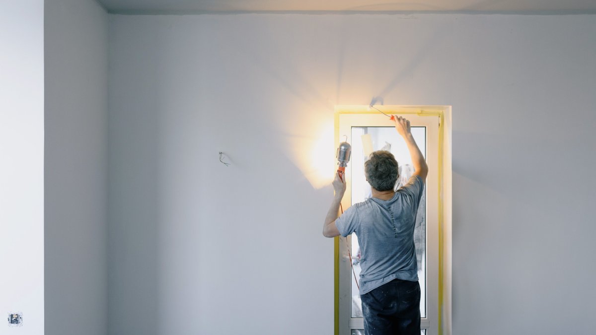 Back view of a worker in work clothes holding a bright lamp while repairing a door at home, representing detailed wall repair work.